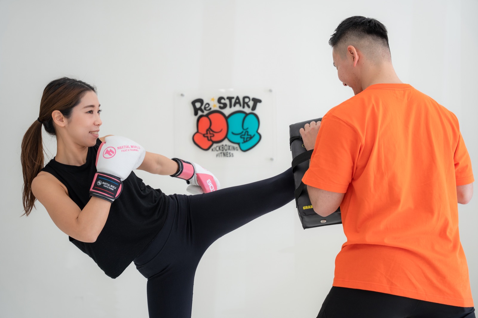 笑顔でミットへキックする女性と、サポートするトレーナー
